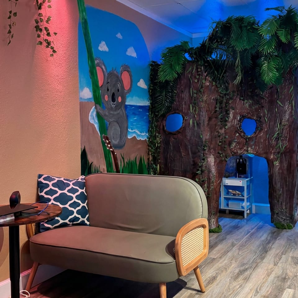 Colorful restaurant lounge with a couch, tropical tree mural, koala wall painting, and illuminated liquor display.