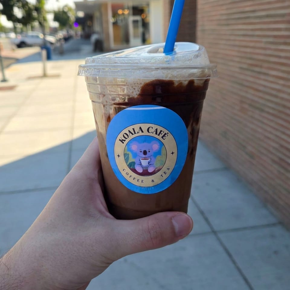 Hand holding an iced coffee from Koala Café on a sunny sidewalk beside a brick wall.