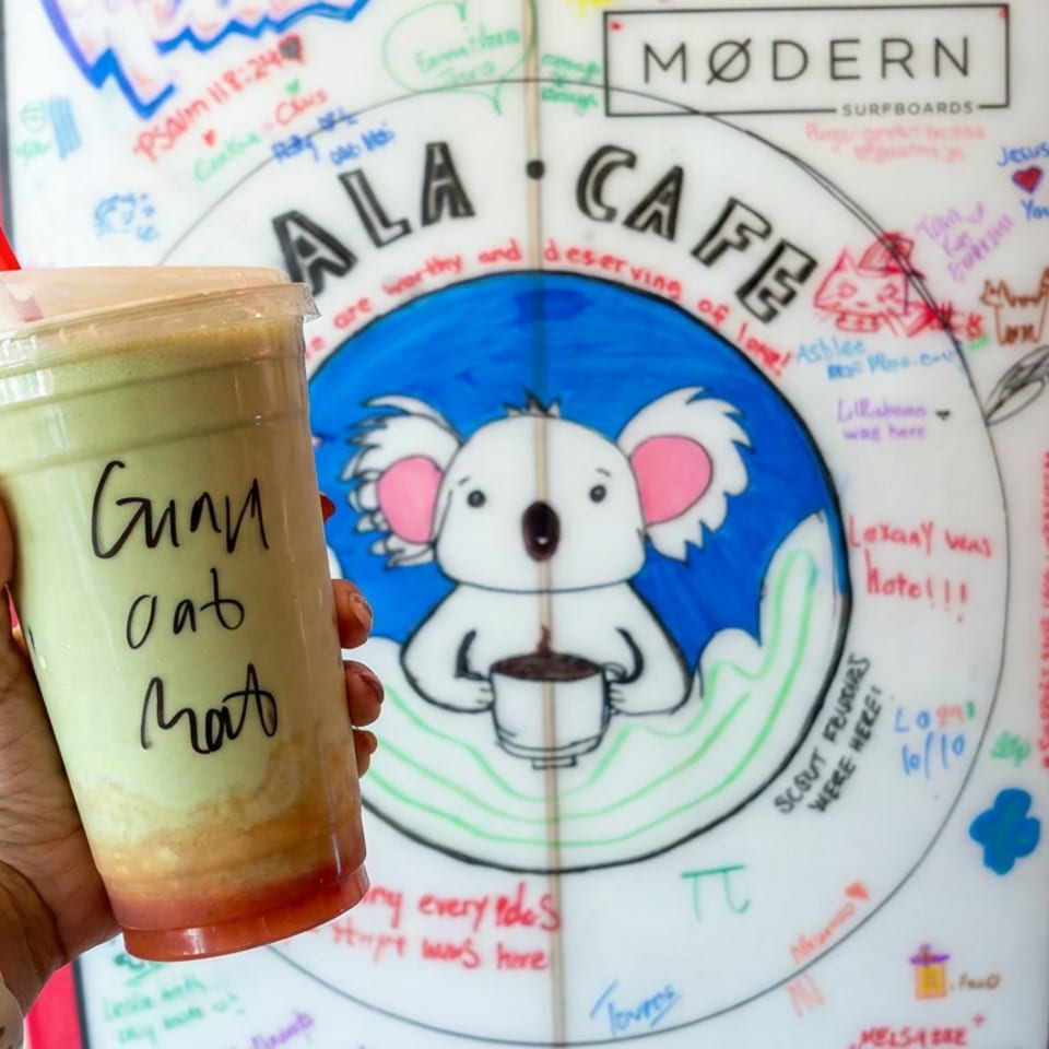 Hand holding a layered matcha drink with red syrup, in front of a colorful koala mural covered with handwritten messages.