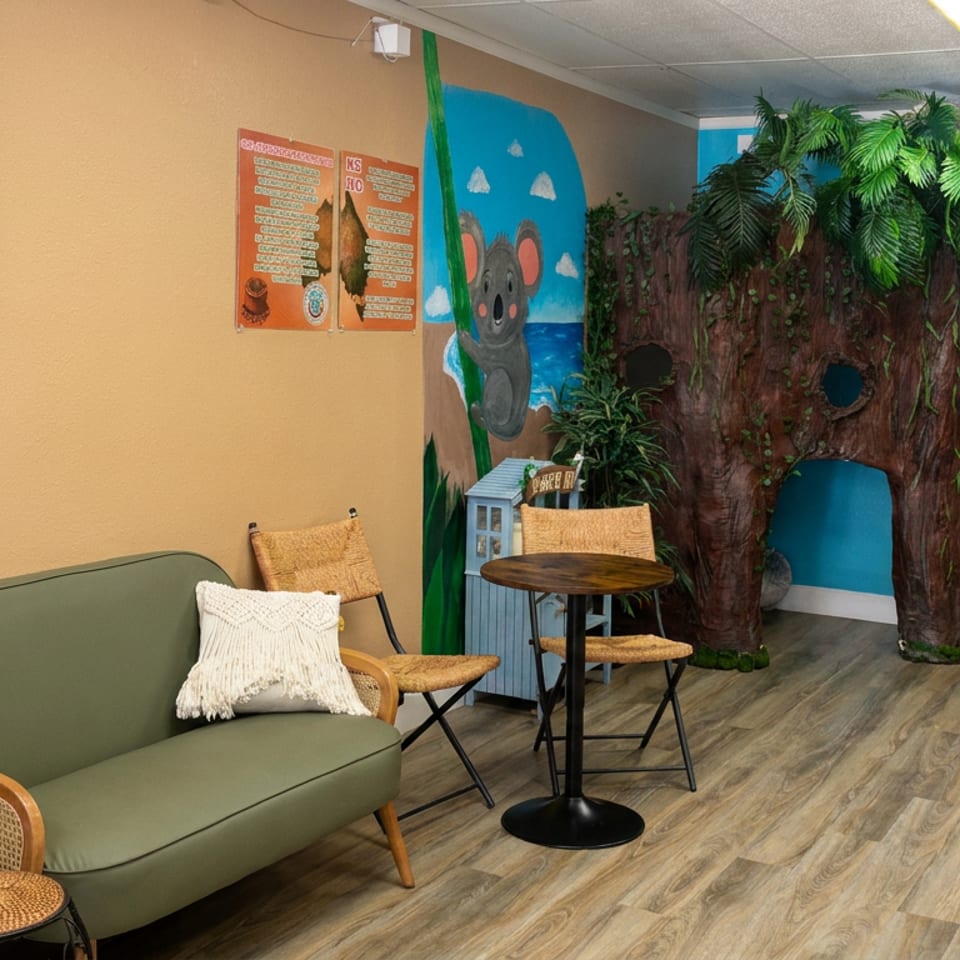 Colorful restaurant interior with ocean artwork, cozy seating, tropical plants, koala mural, tree-themed play area, and hanging packaged treats.